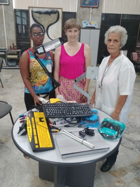 A medium shot shows three women standing behind a round table filled with assistive technology devices. The woman on the left has dark skin, wears glasses, and a colorful tank top. The woman in the center has light skin and short brown hair; she wears a bright pink tank top and a floral skirt. The woman on the right has light skin and short white hair; she wears a white lab coat over a light-colored top. The table holds various items including a yellow keyboard with large black keys, a standard black computer keyboard, a silver laptop, a silver pen, and a teal-colored device with a clear cover. A flexible arm lamp is positioned over the table, illuminating the devices. The women appear to be looking at the camera. The background shows a room with light-colored walls, a tiled floor, artwork on the walls, and some furniture, suggesting an educational or workshop setting.