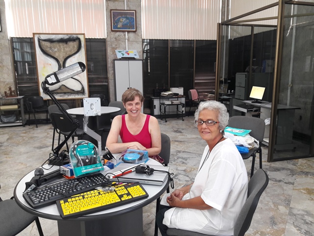 A medium shot shows two women seated at a round table in a well-lit room. The woman on the left appears to be of light skin tone with short brown hair. She is smiling and wearing a bright pink tank top. The woman on the right also appears to be of light skin tone with short white hair and glasses. She is wearing a white top and a lanyard with a badge. She is looking towards the camera with a slight smile.
The table between them is covered with various assistive technology devices, including a yellow keyboard with large black keys, a standard black computer keyboard, a silver laptop, and a teal-colored device in a clear plastic case with blue packaging. A flexible arm lamp is positioned over the table, illuminating the items.
The background shows a room with light-colored walls, large windows with blinds, a tiled floor, and other tables and chairs. There is also artwork on the walls and a glass-enclosed area with computer equipment. The setting suggests a workshop, educational, or resource center focused on assistive technology.