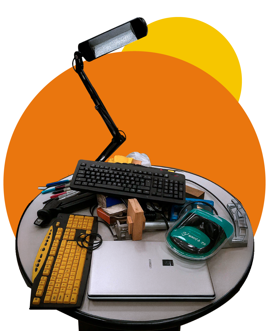 An aerial view of a cluttered, round desk. In the center, there is a silver laptop topped with a black keyboard and a yellow and black keyboard. Above the laptop, a black desk lamp is lit, pointing downwards. Around the main items, various small objects like pens, a green and black mouse, boxes, and wooden pieces are seen piled up. The background is gray with two large, overlapping circles in shades of orange and yellow in the upper right.