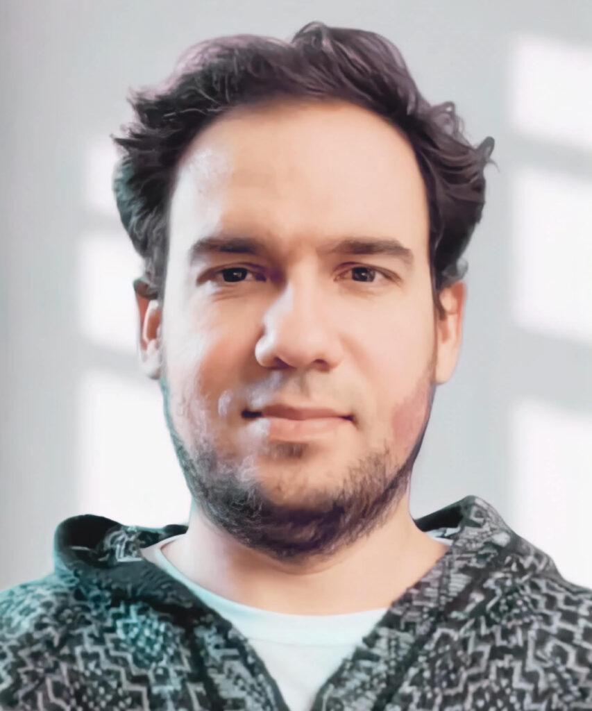 A portrait of a young man with dark hair and stubble, looking directly at the camera. He is wearing a knit hoodie with a geometric black and white pattern. The background is a white wall with a window's light and shadow pattern.