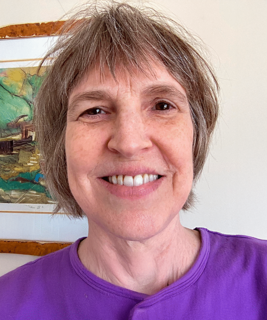 A close-up, smiling portrait of a middle-aged woman with short brown hair, wearing a purple t-shirt. Behind her, partially visible, is a painting of a landscape in green and yellow tones and a gold wooden frame.