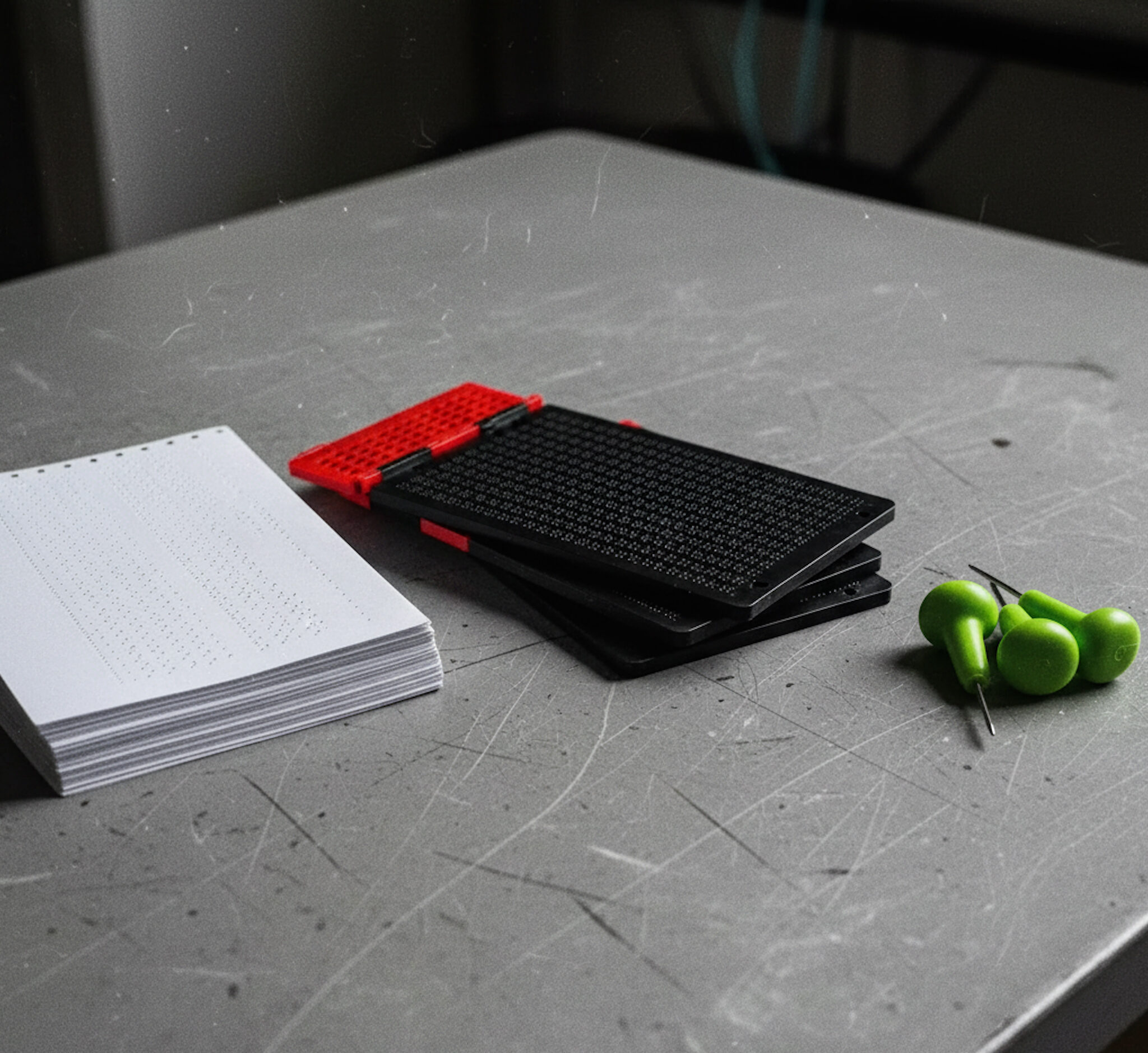 A close-up view of a textured gray table displaying Braille writing tools. On the left, there is a stack of Braille paper with perforated dots. In the center, there is a black and red Braille slate and stylus. On the right, there are three individual Braille styluses with bright green handles.